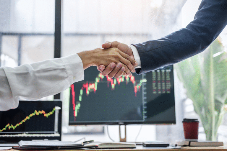 Two professionals shaking hands in front of stock market graphs after completing a business deal or acquisition