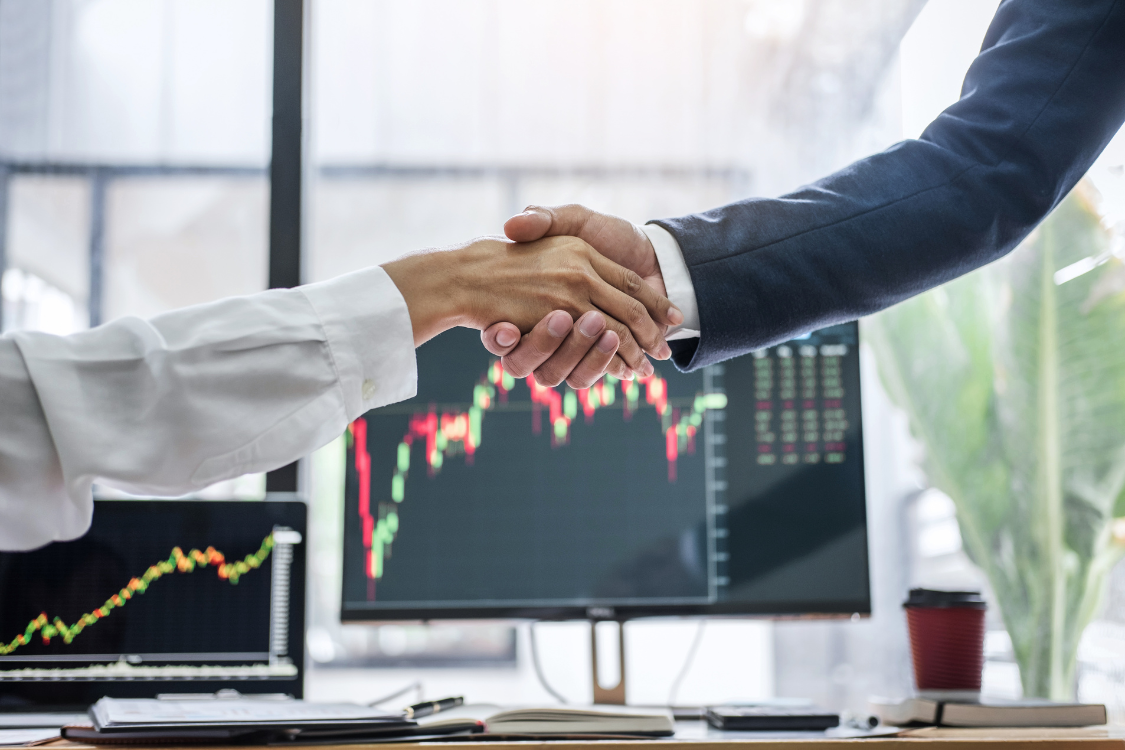 Two professionals shaking hands in front of stock market graphs after completing a business deal or acquisition