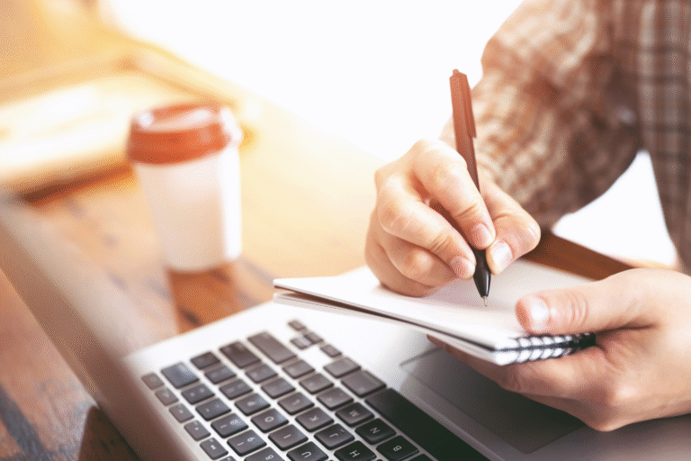 Man writing notes with pen near laptop and coffee cup, reviewing business broker information