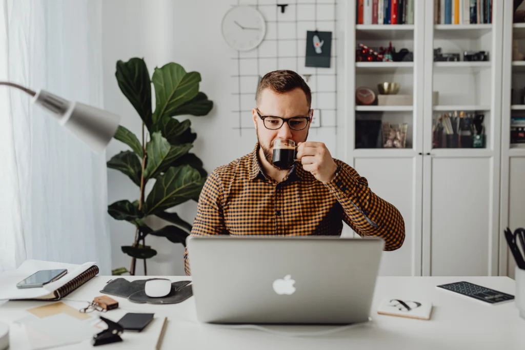 A financial broker or advisor reviewing documents on a laptop while drinking coffee, preparing for a business sale transaction outlined in an engagement letter or broker agreement.