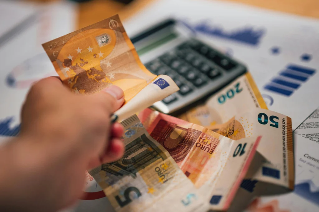 Hand counting Euro currency notes with a calculator in the background, illustrating the calculation of broker fees defined in an engagement letter.