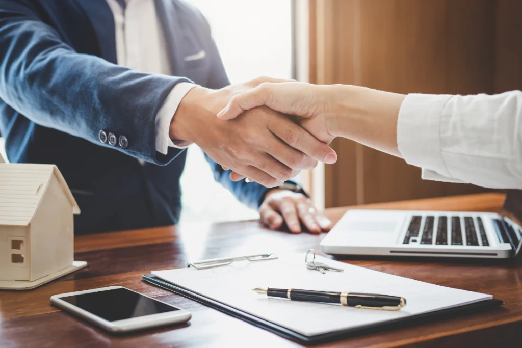 A professional handshake over legal documents and a model house, representing an agreement or the end of a partnership defined by an engagement letter and its tail period.