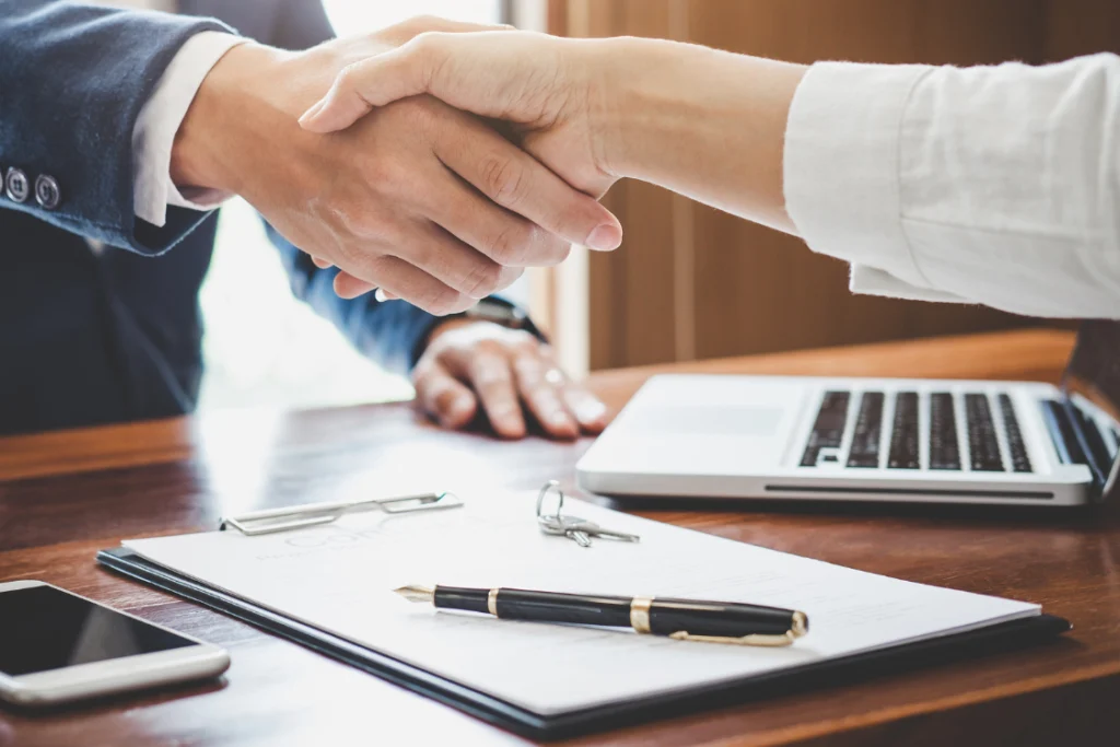 A close-up of a handshake over a signed document with a pen and keys, representing the formal execution of a broker agreement or engagement letter in a business sale.