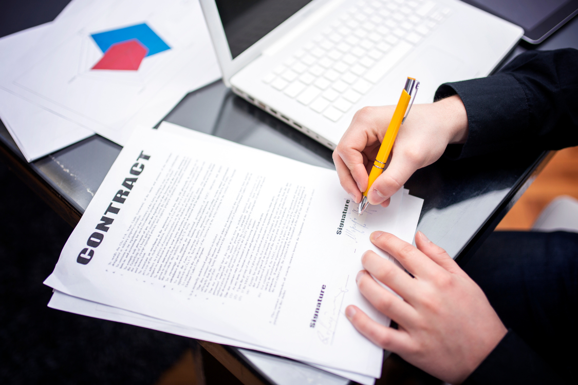 High-angle shot of a person signing a business sale contract with a yellow pen, detailing the signature section of an exclusivity clause engagement letter.