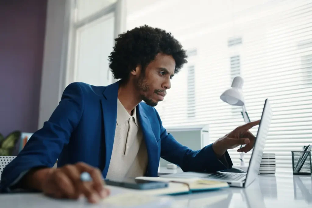 A male financial analyst in a blue suit intently points at his laptop screen while reviewing data for a private business valuation or complex financial modeling.