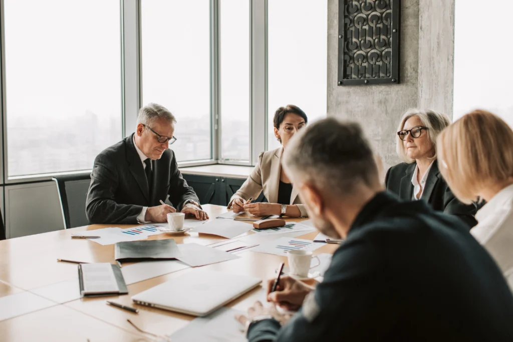 A group of professionals in suits and business attire seated around a conference table, signifying a meeting to align the team and broker on the business listing launch date and strategy.