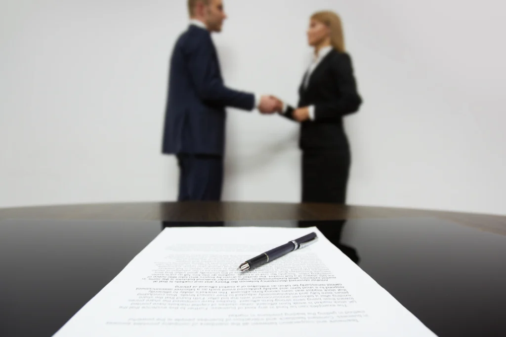Focus on a pen resting on a sample engagement letter for business sale contract, with two business brokers shaking hands blurred in the background, symbolizing a closed deal.