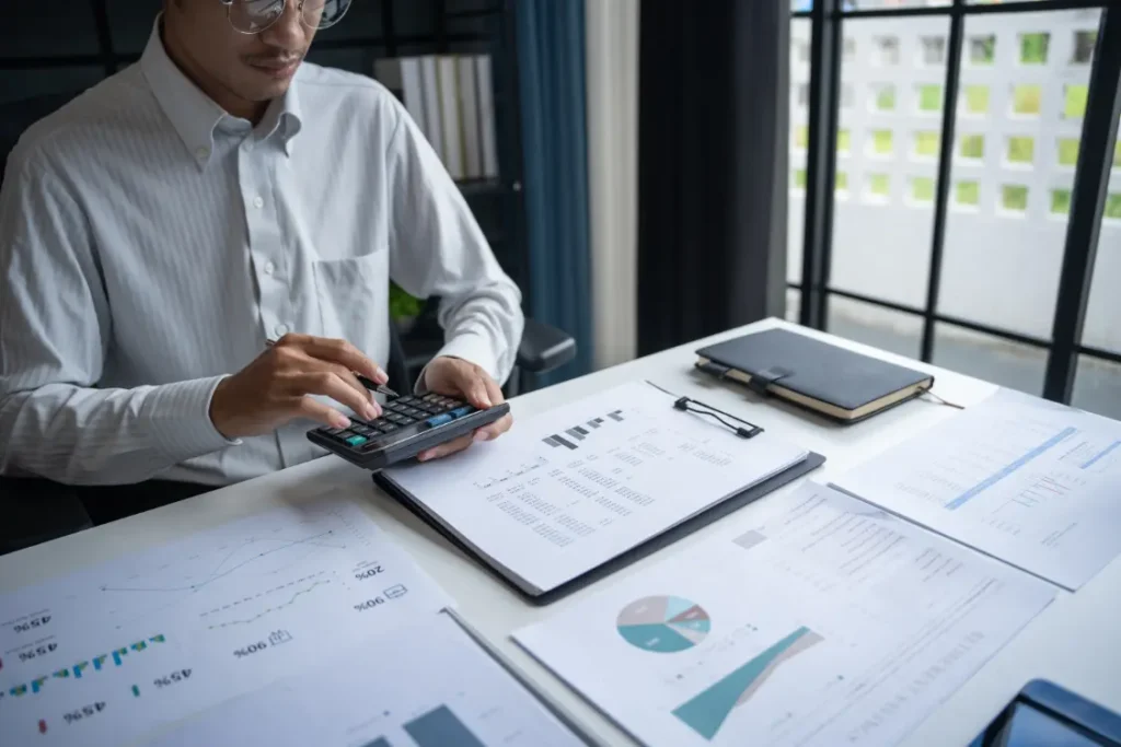 An analyst using a calculator with financial statements and charts spread across the desk, showing the detailed calculation work required for a private business valuation.