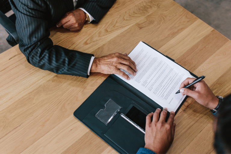 An overhead shot of two individuals signing a sample engagement letter for business sale on a wooden table, emphasizing the final step of agreement.