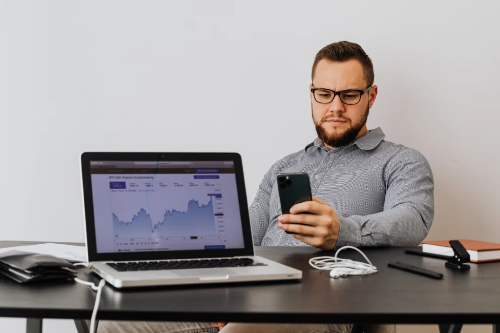 A focused man looking at his phone while a laptop displays a chart, representing a business owner or broker reviewing Key Performance Indicators (KPIs) after the business listing launch date.