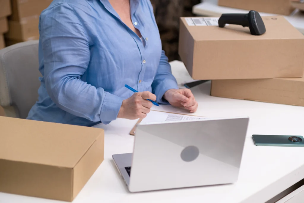 A sales rep or business owner tracking leads on a laptop and paper near shipping boxes, symbolizing the use of tools to manage and fulfill qualified sales.