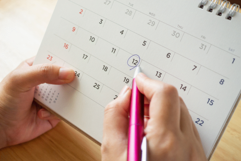 A hand with a pink pen circling a date (the 12th) on a paper calendar, symbolizing the action of setting and confirming the business listing launch date.