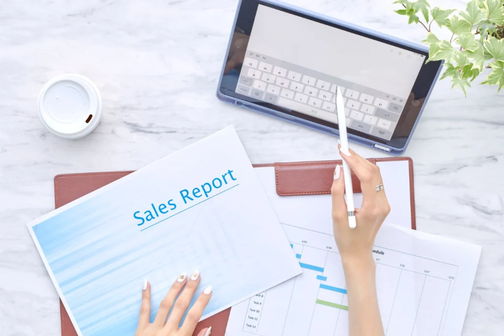 A person's hands near a tablet and coffee, working on a document labeled "Sales Report," which relates to setting up a business sale KPI tracker template.