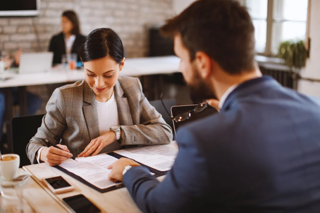 A woman in a business suit signing a sample engagement letter for business sale document, with a male business broker pointing to a section.