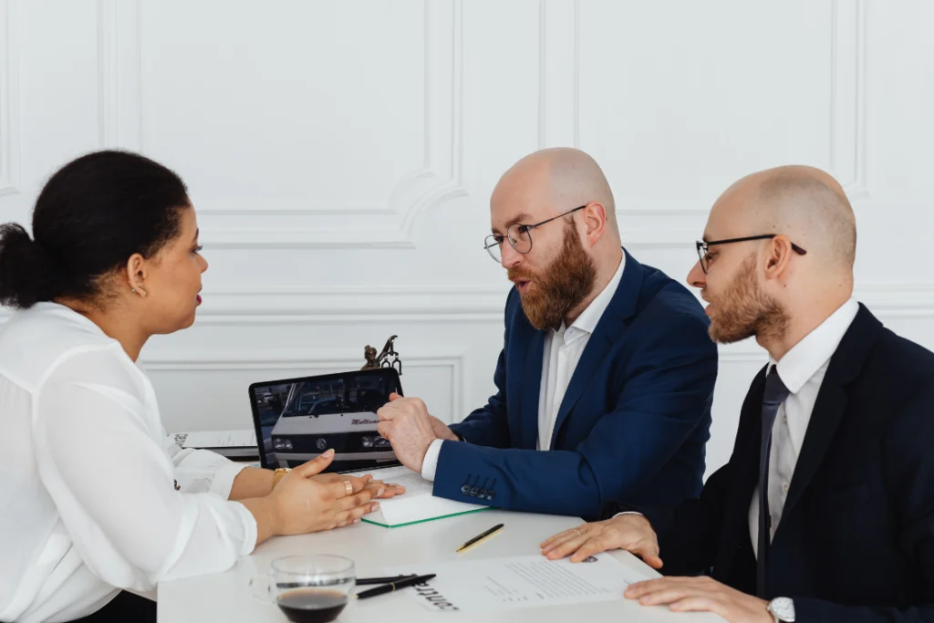 Three business professionals discussing an offer or contract, with one pointing to a laptop screen, using KPIs from the business sale KPI tracker template to guide the conversation.
