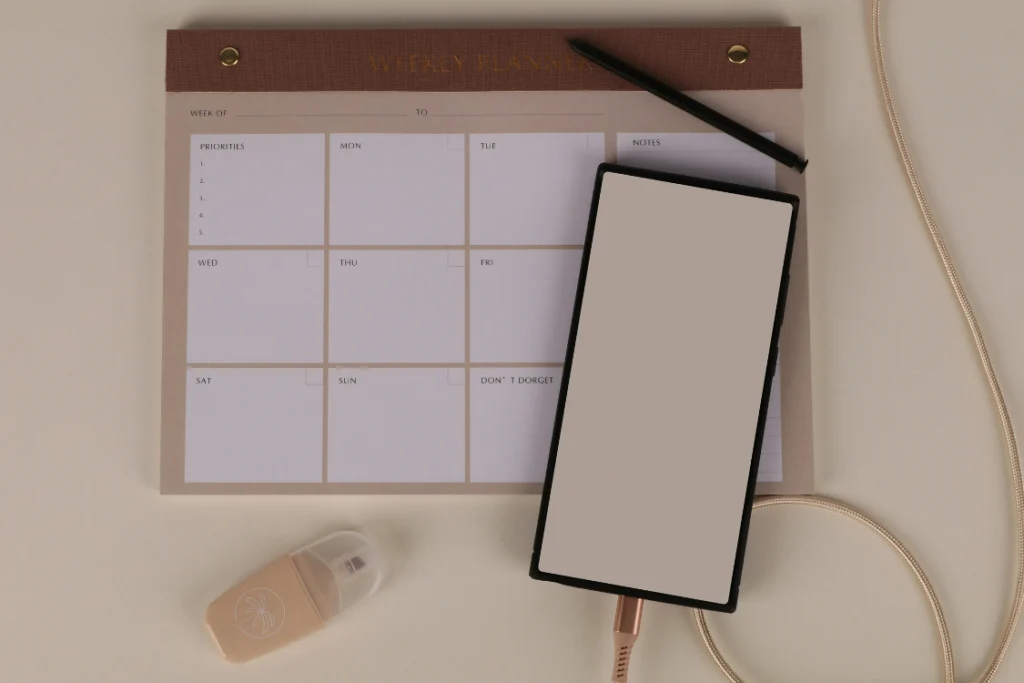 A weekly planner pad with sections for priorities and days of the week next to a smartphone, illustrating the use of a schedule to manage tasks leading up to the business listing launch date.