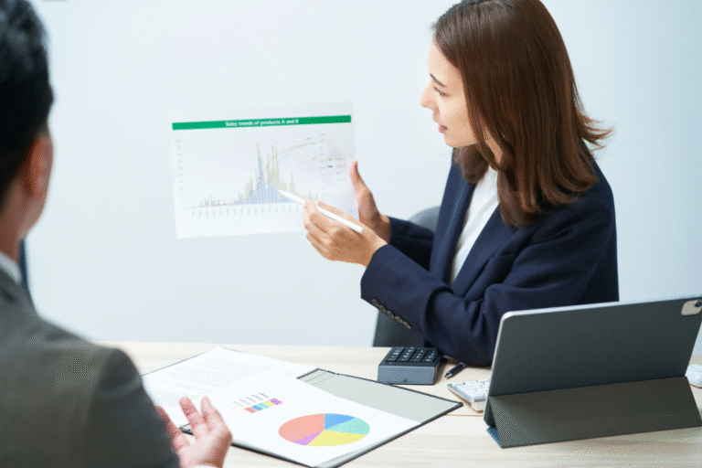 A woman in a business suit pointing to a graph showing sales trends and KPI data to a colleague, discussing the progress of a business sale KPI tracker template.