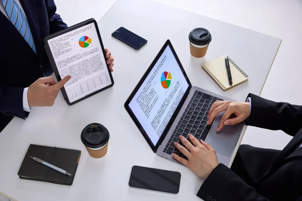 Overhead view of two professionals reviewing a business valuation report with pie charts on a laptop and tablet, emphasizing the role of a business valuation expert in due diligence.
