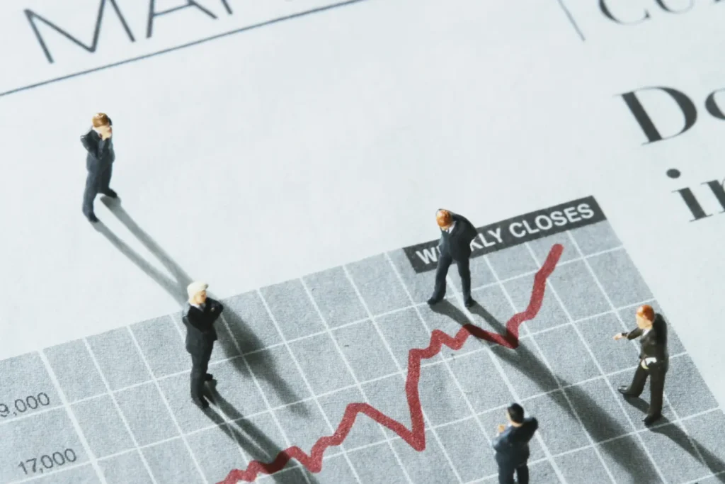 Miniature business figures standing on a financial newspaper and an upward-trending red line graph, symbolizing market trends and the impact of a business valuation on economic value.
