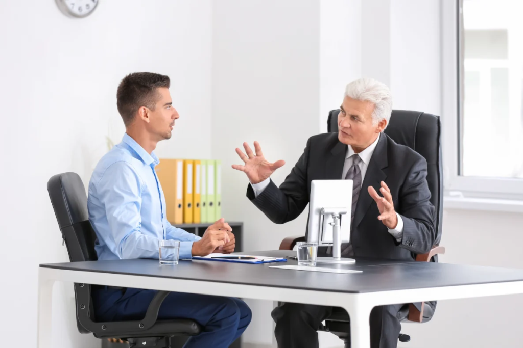 Two individuals are seated at a desk in an office, engaged in a conversation.