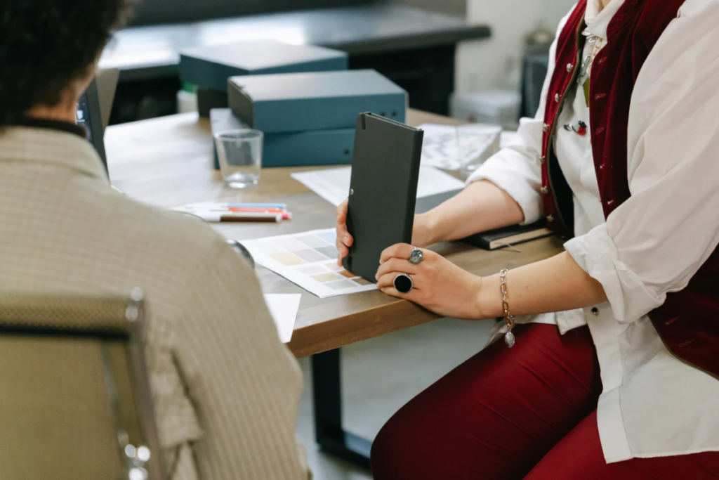 Two people are sitting at a desk, one is holding a black folder and looking at color samples on the desk.