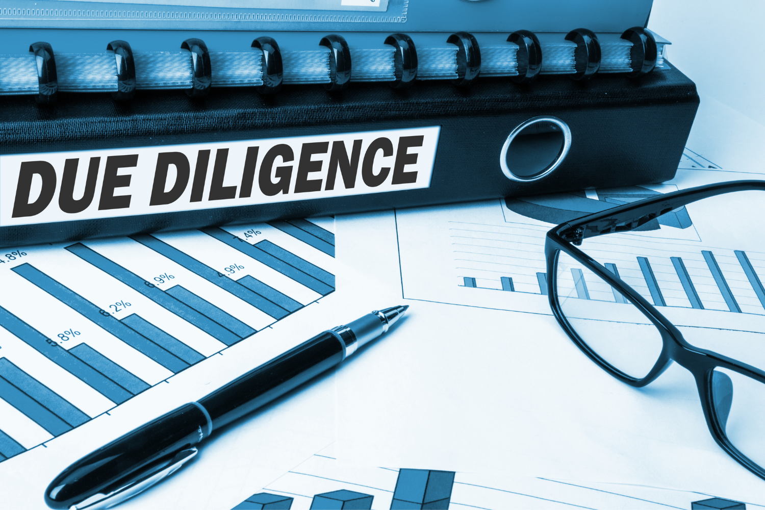 A binder labeled "DUE DILIGENCE" sits on top of financial charts next to a pen and eyeglasses.