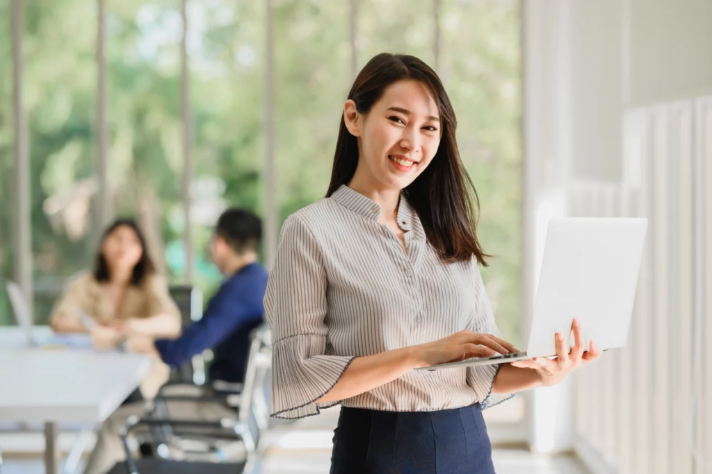 A smiling person holds a laptop in a bright office with colleagues blurred in the background.