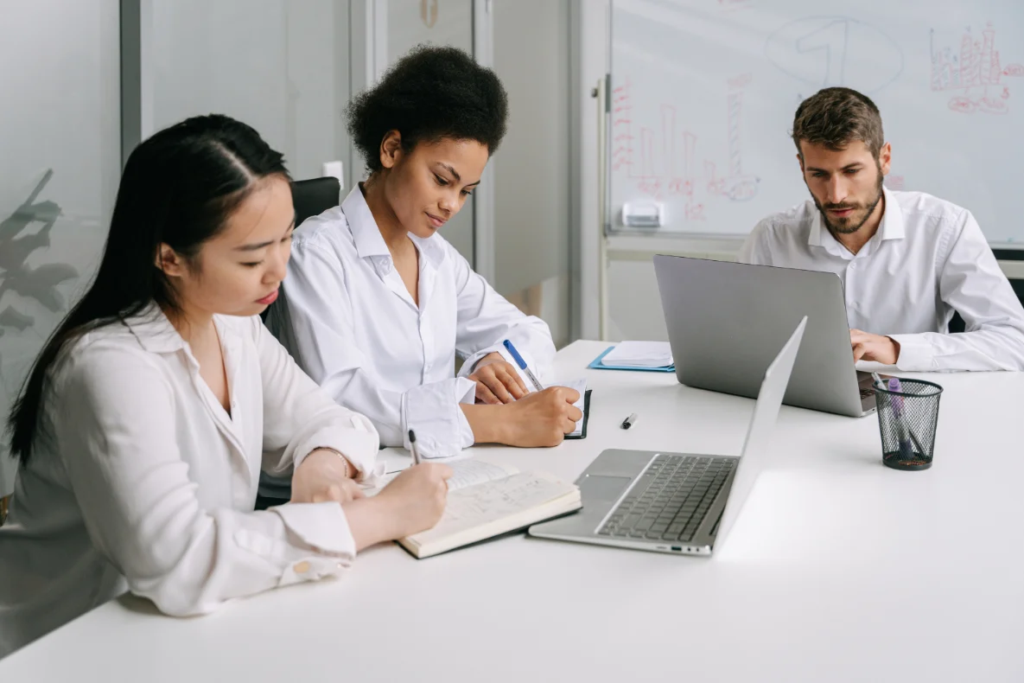 Three people in white shirts are sitting at a white table with laptops and notebooks.