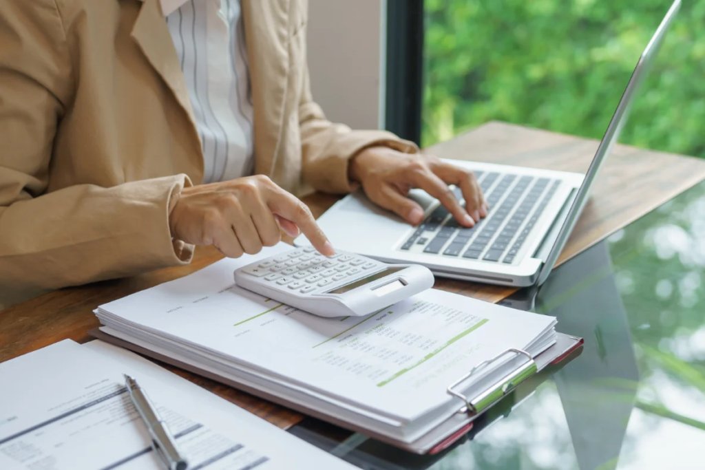 A person in a tan blazer uses a calculator and types on a laptop while reviewing documents on a clipboard on a table.