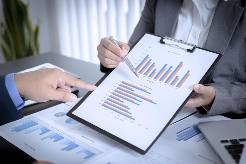 Hands pointing at bar charts and graphs on a clipboard and desk, suggesting a business meeting or financial review.