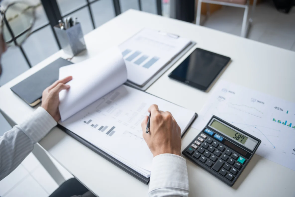A person's hands are shown over a white desk with various documents, a tablet, and a calculator. One hand is turning a page of a document, while the other holds a pen and appears to be writing on a notepad.
