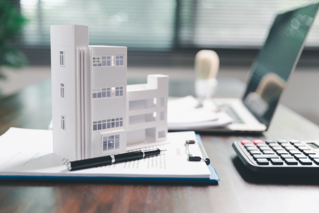 A white architectural model of a building sits on a desk next to a laptop, calculator, and documents with a pen on a clipboard.