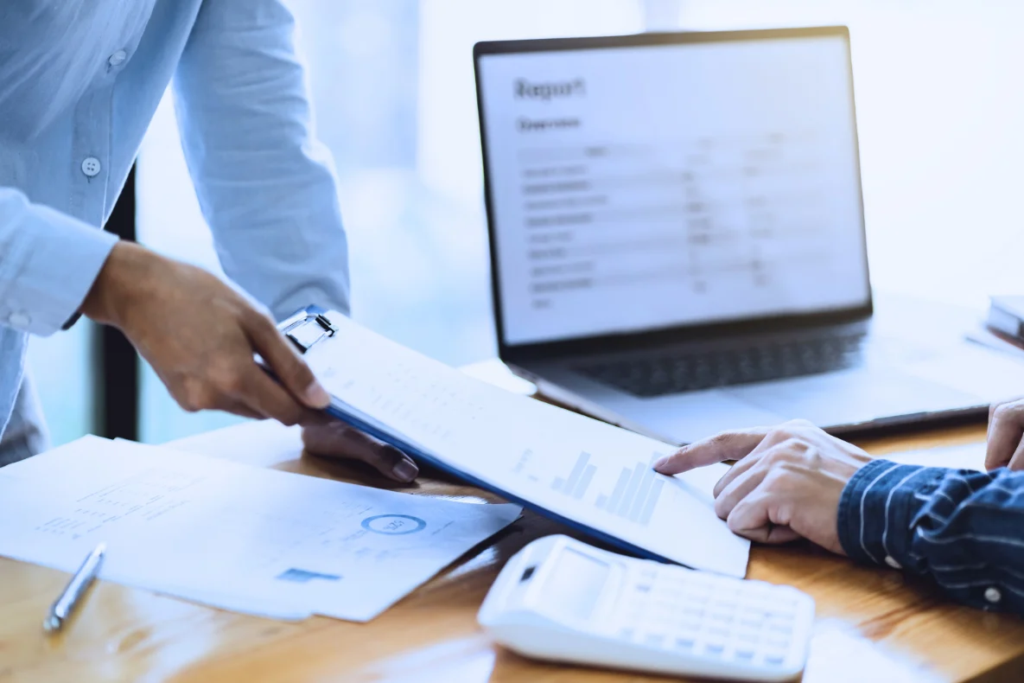 A person hands a document on a clipboard to another person who is pointing at it, with a laptop and calculator on the table.