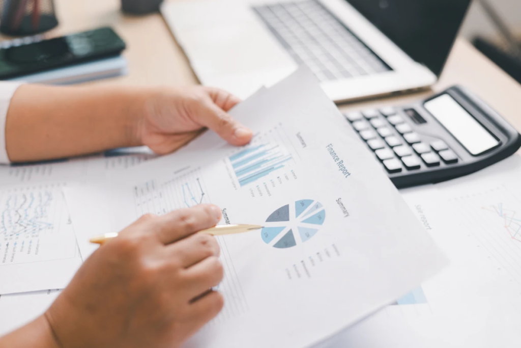 A person's hands holding a financial report with charts and a pie graph. A calculator and laptop are visible in the background.