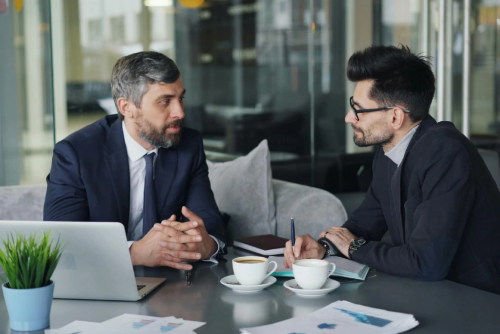 Two individuals in suits are seated at a table with laptops, coffee, and papers, engaged in a discussion.