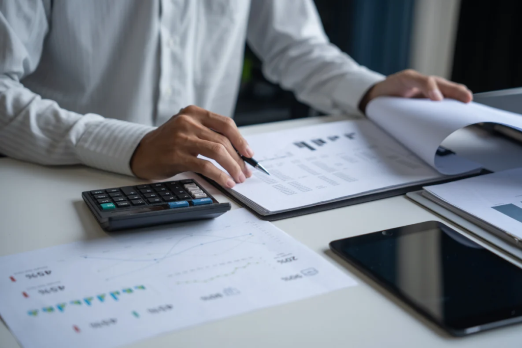 A person in a white shirt is reviewing documents at a desk with a calculator, a tablet, and various charts and graphs.