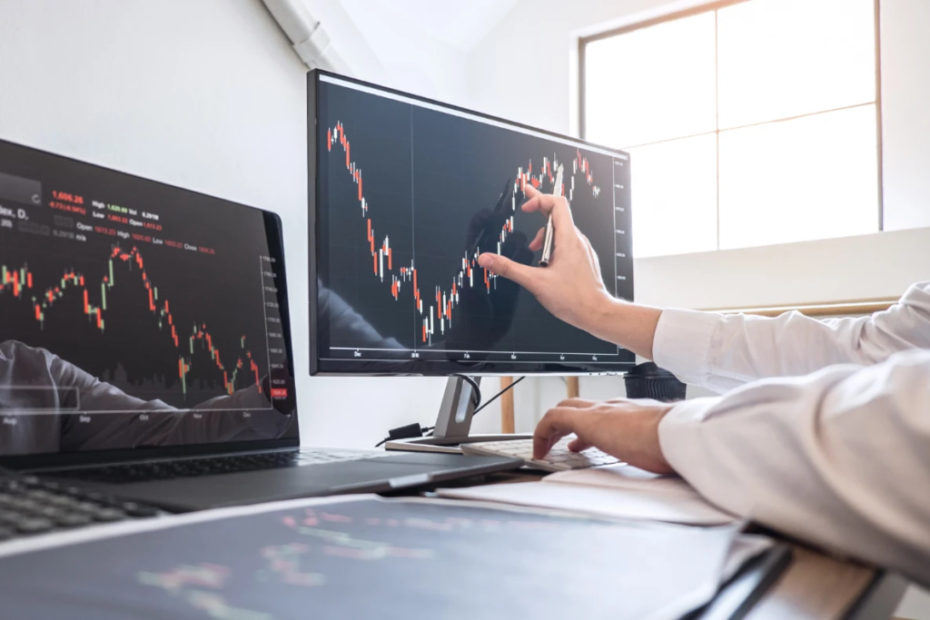 A person points to a stock market chart on a monitor while typing on a keyboard, with another monitor and laptop displaying similar charts.