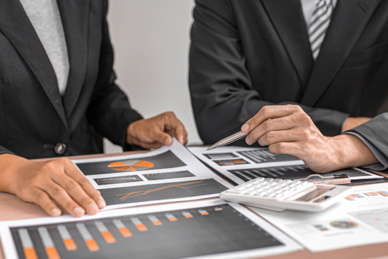 Two business professionals review financial charts, reports, and a calculator while discussing how to price a business for sale accurately.