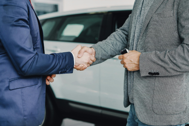 Two businesspeople shaking hands beside a car during a sale or deal negotiation.